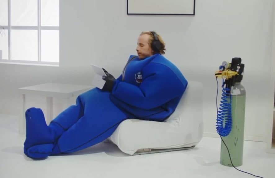 A man in the BodyStream suit relaxing in a chair with a tablet, the CO2 tank and regulator beside him